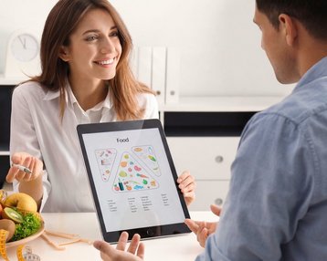 Fernstudium Bachelor Ernährungswissenschaft und Prävention | SRH Fernhochschule Eine lächelnde Frau mit lockigem Haar sitzt an einer Küchentheke, schreibt in ein Notizbuch und umgeben von frischem Gemüse und Obst.