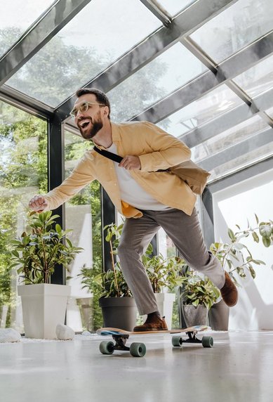 Mann in Anzug fährt auf einem Skateboard in einem modernen Raum mit großen Fenstern und Pflanzen.