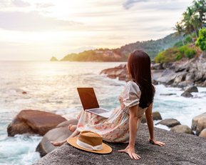 Eine Frau sitzt auf einem Felsen am Meer, mit einem Laptop auf den Knien und einem Hut neben sich, während die Sonne untergeht.