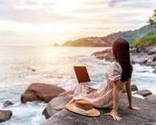 Eine Frau sitzt auf einem Felsen am Meer, mit einem Laptop auf den Knien und einem Hut neben sich, während die Sonne untergeht.