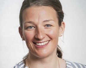 Eine lächelnde Frau mit blonden Haaren trägt ein gestreiftes Hemd. Sie hat Ohrringe und schaut direkt in die Kamera.