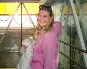 Eine Person mit blonden Haaren trägt einen rosa Hoodie und hält eine Jacke, während sie in einem modernen Gebäude steht.