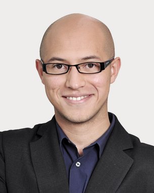 Mann mit Glatze und Brille, trägt einen schwarzen Anzug und ein dunkles Hemd, lächelt freundlich und steht mit verschränkten Armen.