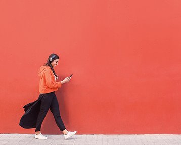 Eine Person mit Kopfhörern trägt einen orangefarbenen Hoodie und schwarze Hosen, während sie an einer roten Wand entlanggeht und ein Smartphone in der Hand hält.