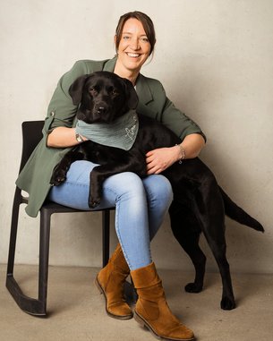 Eine Frau sitzt auf einem Stuhl und hält einen schwarzen Labrador, der ein Halstuch trägt. Sie lächelt in die Kamera.
