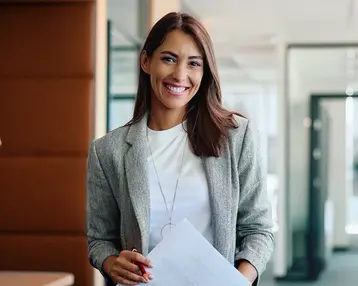 Eine lächelnde Frau in einem modernen Büro hält ein Dokument in der Hand. Sie trägt einen grauen Blazer und hat lange, braune Haare.