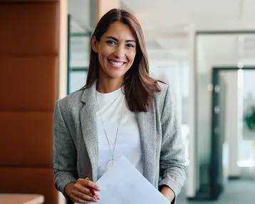 Eine lächelnde Frau in einem modernen Büro hält ein Dokument in der Hand. Sie trägt einen grauen Blazer und hat lange, braune Haare.