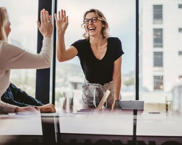 Eine Frau mit Brille lächelt und gibt einer anderen Person einen High-Five in einem modernen Besprechungsraum. Im Hintergrund sind große Fenster und einige Gläser sichtbar.