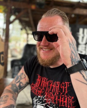 Mann mit Bart und Sonnenbrille, trägt ein schwarzes T-Shirt mit rotem Aufdruck, sitzt lächelnd an einem Tisch im Freien.