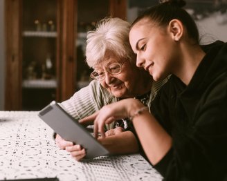 Eine ältere Frau und eine junge Frau sitzen an einem Tisch und schauen gemeinsam auf ein Tablet. Beide lächeln, während sie sich auf den Bildschirm konzentrieren.