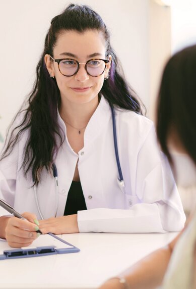 Eine Frau in einem weißen Kittel sitzt an einem Tisch und spricht mit einer Patientin, während sie Notizen macht.