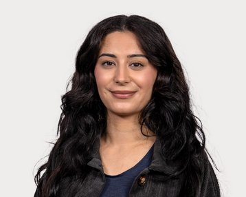 Eine Frau mit langen, lockigen, schwarzen Haaren lächelt in die Kamera. Sie trägt ein schwarzes Oberteil und hat einen neutralen Hintergrund.