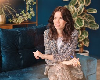 Eine Frau mit lockigem, braunem Haar sitzt auf einem blauen Sofa, trägt einen karierten Anzug und hält einen Stift. Sie schaut nachdenklich und ist in einem modernen Raum mit Pflanzen und dekorativen Elementen.