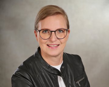 Eine Person mit blonden Haaren und Brille trägt eine schwarze Lederjacke und ein weißes T-Shirt. Sie lächelt und sitzt vor einem neutralen Hintergrund.