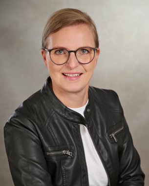 Eine Person mit blonden Haaren und Brille trägt eine schwarze Lederjacke und ein weißes T-Shirt. Sie lächelt und sitzt vor einem neutralen Hintergrund.
