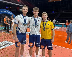 Drei Sportler stehen auf dem Spielfeld, tragen Medaillen und feiern ihren Sieg. Der Boden ist mit Konfetti bedeckt.