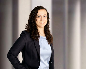 Eine lächelnde Frau mit lockigem, braunem Haar trägt einen schwarzen Blazer und ein hellblaues Oberteil. Sie steht mit einer Hand an der Hüfte vor einem neutralen Hintergrund.