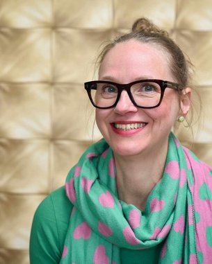 Eine Frau mit Brille und einem bunten Schal mit Herzen lächelt vor einem goldfarbenen, strukturierten Hintergrund.