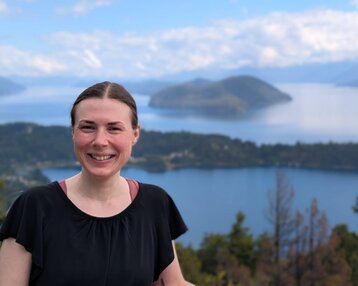 Eine Frau mit langen Haaren lächelt vor einer malerischen Seenlandschaft mit Bergen im Hintergrund.