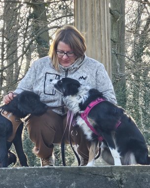 Eine Frau sitzt auf einer Mauer und streichelt zwei Hunde, während sie in einem Waldgebiet umgeben von Bäumen sind.
