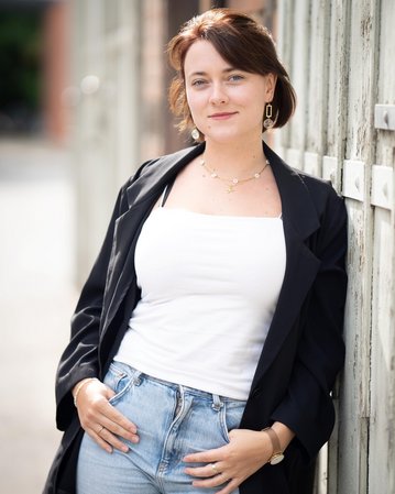 Eine Person mit kurzen, braunen Haaren trägt ein schwarzes Blazer und ein weißes Top, während sie an einer Wand lehnt.